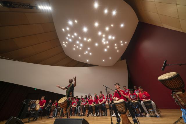 Des jeunes portant un t-shirt jouant du djembé sur scène avec leur professeur au premier plan