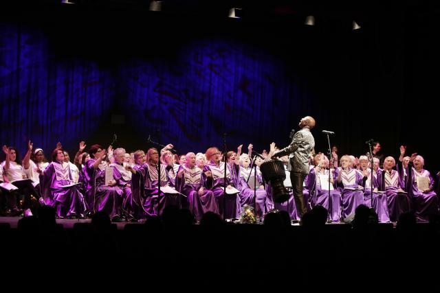 Une large chorale de personnes en robe violette chantant sur scène