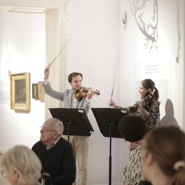 Deux violinistes jouant devant un groupe de personnes assises dans un musée