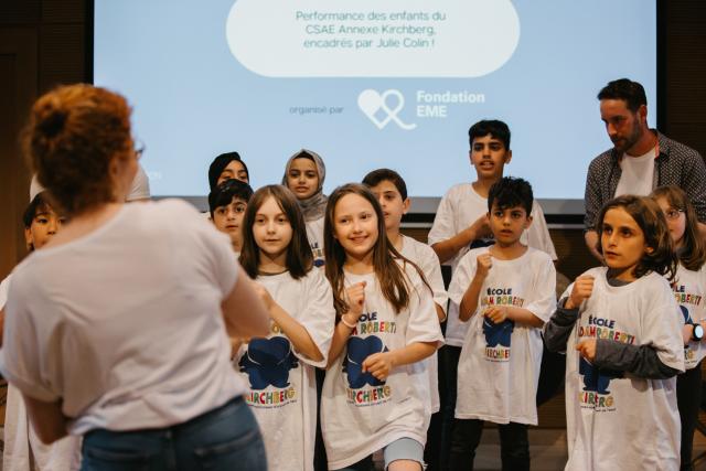 Un groupe d'enfants portant le même t-shirt blanc sur scène devant leur professeur de dos