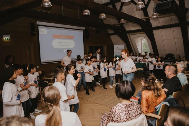 Un groupe d'enfants portant le même t-shirt blanc sur scène devant leur professeur