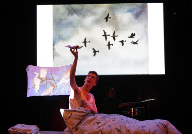 Un homme sur scène allongé dans un lit et jouant avec une projection d'oiseau