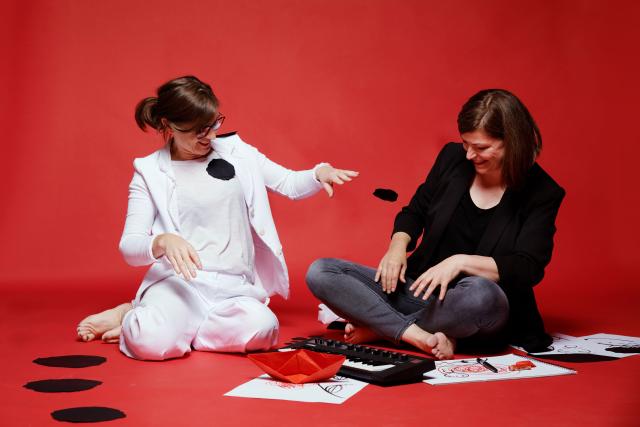 Deux femmes assises au sol entourées de dessins et d'un clavier, sur un fond rouge