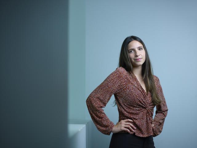 Une fille avec des cheveux bruns qui sourit. Elle porte une blouse rouge et pose avec les mains sur sa taille. 